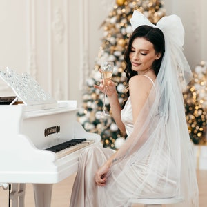 May include: A woman in a white dress and veil sits at a white grand piano, holding a glass of champagne. A decorated Christmas tree is in the background.