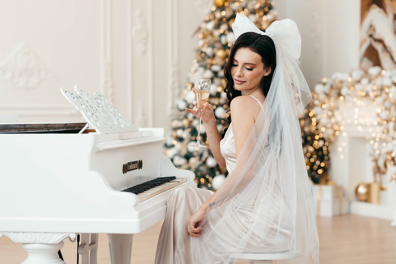 May include: A woman in a white dress and veil sits at a white grand piano, holding a glass of champagne. A decorated Christmas tree is in the background.