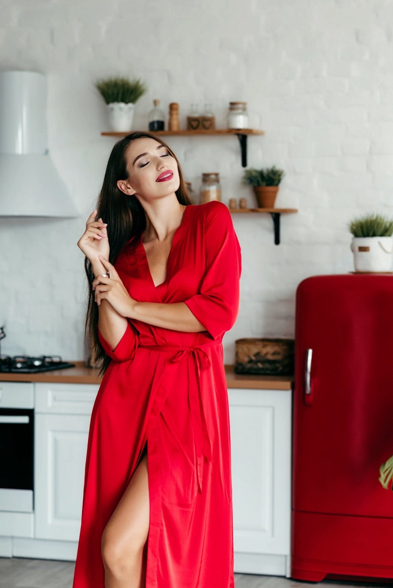 May include: A woman wearing a red satin dressing gown stands in a kitchen with a red fridge in the background. She is smiling and looking upwards.
