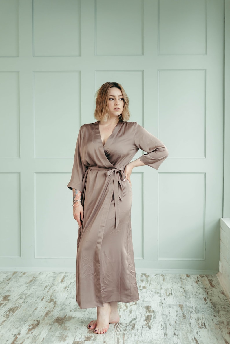 May include: A woman wearing a long, brown satin robe with a wrap style and a V-neck. The robe has a sash tied at the waist.