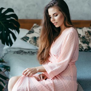 May include: A woman in a pink polka dot robe sits on a bed with a white and green patterned pillow behind her.
