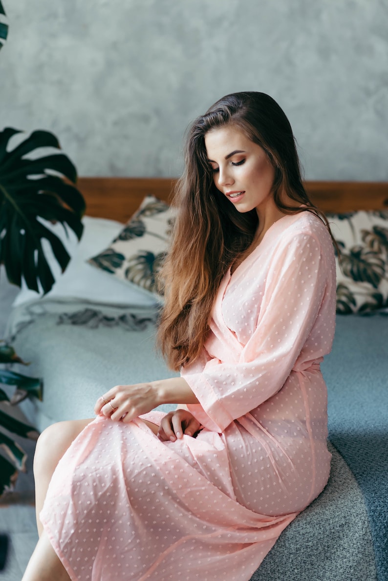 May include: A woman in a pink polka dot robe sits on a bed with a white and green patterned pillow behind her.