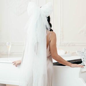 May include: A woman wearing a white veil with a large bow on top, standing behind a white grand piano. She is wearing a light brown slip dress.