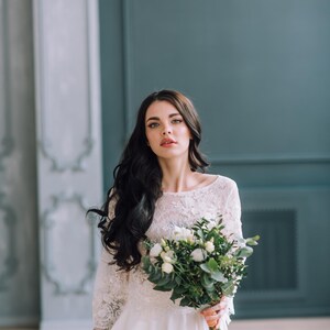 Op de afbeelding: Een witte jurk met een kanten lijfje met driekwartmouwen en een zwierige rok. Het kledingstuk wordt gecombineerd met een boeket witte bloemen en groen blad, vastgehouden door een persoon met lang donker haar en een neutrale uitdrukking.