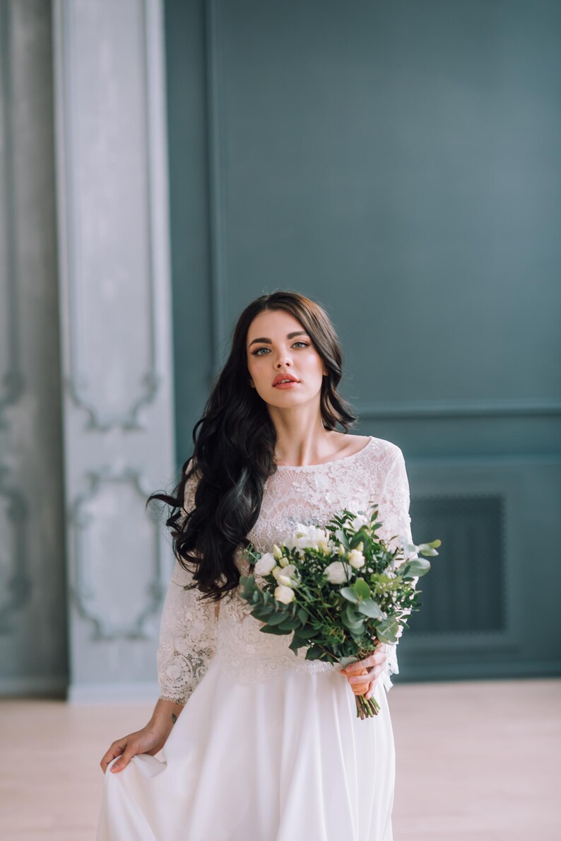 Op de afbeelding: Een witte jurk met een kanten lijfje met driekwartmouwen en een zwierige rok. Het kledingstuk wordt gecombineerd met een boeket witte bloemen en groen blad, vastgehouden door een persoon met lang donker haar en een neutrale uitdrukking.