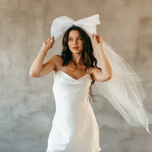 May include: A woman wearing a white satin dress with a large white bow on her head. The bow is attached to a white veil that is flowing behind her.