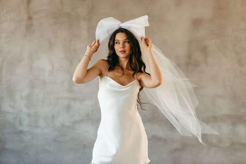 May include: A woman wearing a white satin dress with a large white bow on her head. The bow is attached to a white veil that is flowing behind her.