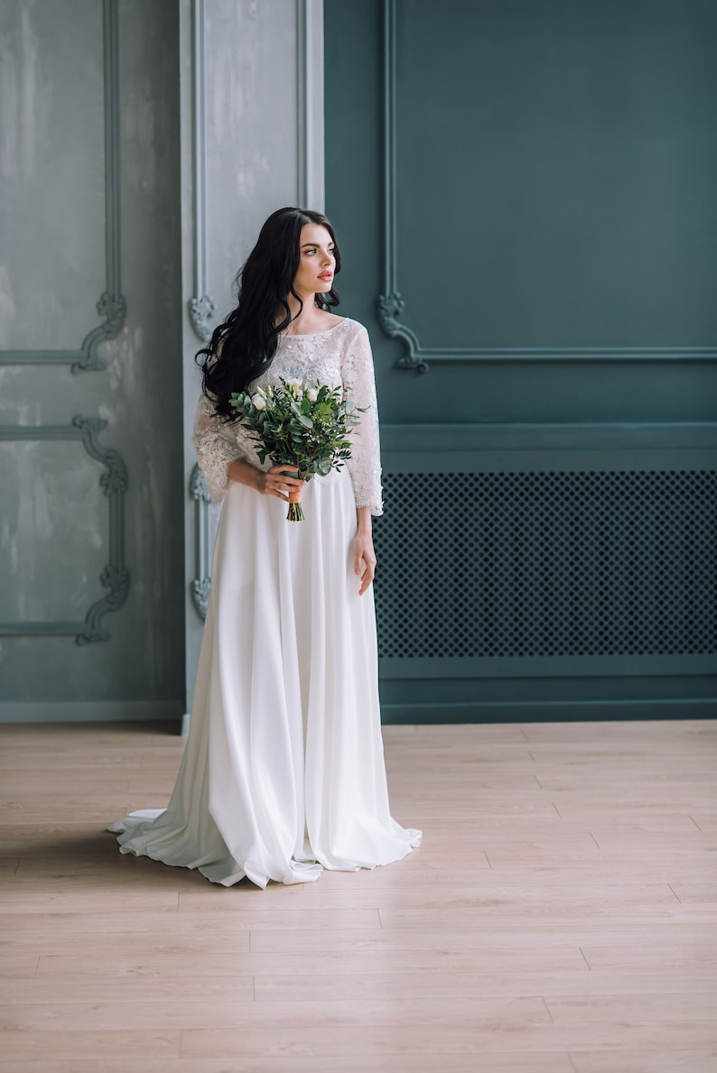 Op de afbeelding: Een vloerlange witte jurk met een kanten lijfje met driekwartmouwen en een effen, zwierige rok. Het kledingstuk wordt gecombineerd met een boeket witte rozen en groen blad, vastgehouden bij de taille in een neutrale binnenruimte.