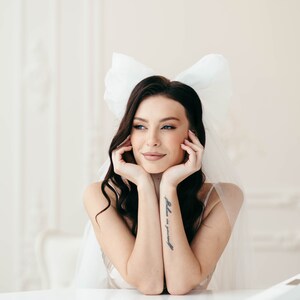 May include: A woman with dark hair wearing a white veil with a large bow on top. She is looking to the side and has her hands resting on her chin. She has a tattoo on her arm that says "believe in yourself".