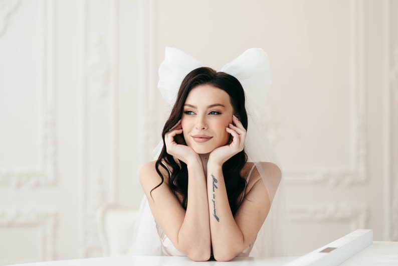 May include: A woman with dark hair wearing a white veil with a large bow on top. She is looking to the side and has her hands resting on her chin. She has a tattoo on her arm that says "believe in yourself".