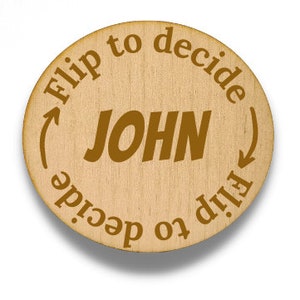May include: A round wooden decision coin with the name "JOHN" in the center. The phrase "Flip to decide" is engraved around the edge with arrows. The coin is light brown with brown text.