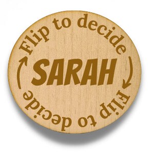 May include: A round wooden decision coin with the name "SARAH" in the center. The text "Flip to decide" is printed around the edge with arrows. The coin is light brown with dark brown text.
