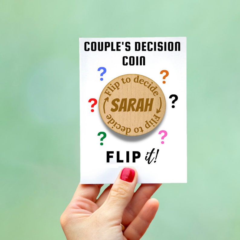 May include: A white card with the text "COUPLE'S DECISION COIN" and "FLIP it!". A wooden coin with "SARAH" in the center is surrounded by "Flip to decide". Colorful question marks surround the coin.