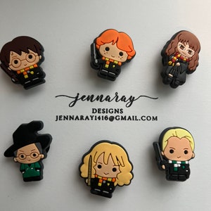 May include: Set of 7 Harry Potter character charms, featuring Harry Potter, Ron Weasley, Hermione Granger, Draco Malfoy, Neville Longbottom, Ginny Weasley, and Luna Lovegood. The charms are made of black, orange, yellow, brown, and green silicone.
