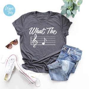 May include: Dark gray t-shirt with the text "What The" above a musical staff with a treble clef and a quarter note. The shirt is paired with blue jeans and white sneakers.