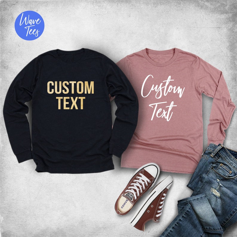 Puede incluir: Dos camisetas de manga larga, una azul marino con texto dorado "CUSTOM TEXT" y otra malva con texto blanco "Custom Text". Zapatillas Converse marrones y vaqueros azules tambi&eacute;n en la imagen. El logo de Wave Tees est&aacute; en la esquina superior izquierda.