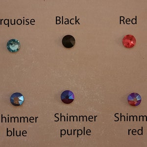 Op de afbeelding: Een selectie ronde, platte strasssteentjes in verschillende kleuren, waaronder wit, turquoise, zwart, rood, groen, transparant, blauw met schittering, paars met schittering en rood met schittering.