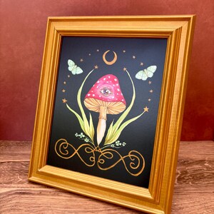 May include: A framed art print featuring a whimsical mushroom design. The artwork showcases a red mushroom with white spots and an eye, set against a black background with a crescent moon, stars, and butterflies. The frame is gold.