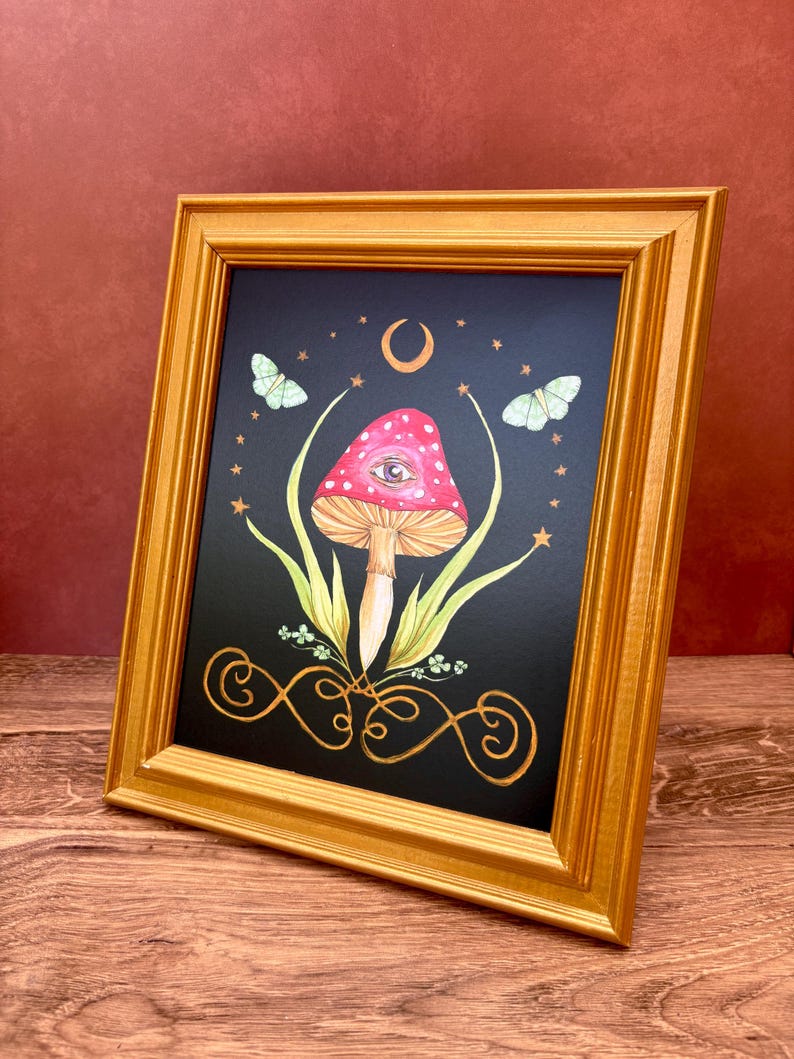 May include: A framed art print featuring a whimsical mushroom design. The artwork showcases a red mushroom with white spots and an eye, set against a black background with a crescent moon, stars, and butterflies. The frame is gold.