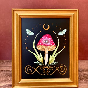 May include: Framed artwork featuring a red mushroom with white spots and an eye design on a black background. The artwork includes butterflies, stars, a crescent moon, and gold decorative elements. The frame is gold.