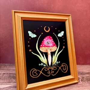 May include: A framed art print featuring a whimsical mushroom design. The mushroom has a red cap with white spots and an eye. The artwork is set against a black background with butterflies, stars, and a crescent moon. The frame is gold.