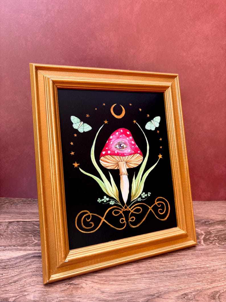 May include: A framed art print featuring a whimsical mushroom design. The mushroom has a red cap with white spots and an eye. The artwork is set against a black background with butterflies, stars, and a crescent moon. The frame is gold.