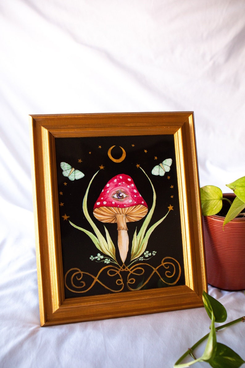 May include: A framed watercolor painting featuring a red mushroom with white spots and an eye design. The artwork is set against a black background with butterflies, stars, and a crescent moon. The frame is gold.
