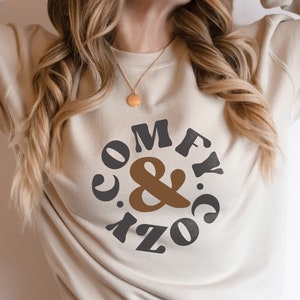 Puede incluir: Una sudadera blanca con un gráfico marrón y negro que dice "Comfy & Cozy" en un círculo.