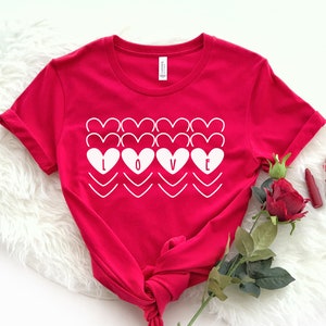 May include: A red t-shirt with the word "LOVE" in white, designed with heart shapes. The shirt is knotted at the bottom. A red rose, lipstick, and a small glass jar with red items are placed near the shirt.