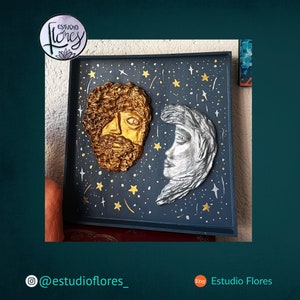 Puede incluir: Una obra de arte enmarcada con un fondo azul oscuro y rostros esculpidos en 3D en oro y plata. El fondo está decorado con estrellas doradas y líneas. El texto "Estudio Flores" es visible en la esquina superior izquierda.