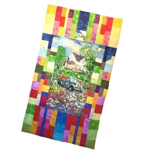May include: A colorful quilt featuring a central image of a farmhouse and a vintage truck filled with produce. The quilt is bordered with vertical rectangles in various colors, including red, yellow, blue, and green. The overall design evokes a pastoral theme.