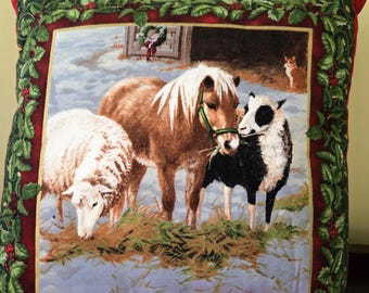 May include: A red, green, and gold pillow with a winter scene featuring a brown and white pony, a white sheep, and a black and white sheep. The animals are standing in a snowy field with a barn in the background.