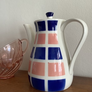 Può includere: Una teiera in ceramica bianca con un motivo geometrico blu e rosa. La teiera ha un beccuccio e un manico. La teiera è appoggiata su una superficie di legno.
