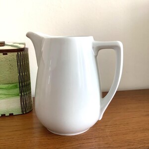 Peut inclure: Une cruche en céramique blanche avec un corps incurvé, un bec verseur et une poignée rectangulaire. La cruche est posée sur une surface en bois, avec un récipient décoratif vert et marron visible à gauche.