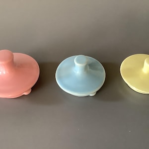 Può includere: Tre coperchi in ceramica dai colori pastello: rosa, azzurro e giallo. Ogni coperchio ha una parte superiore arrotondata con una piccola maniglia. I coperchi sono disposti in fila su uno sfondo grigio.