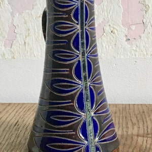 Könnte beinhalten: Eine braune Keramikkanne mit einem blauen und goldenen Blumendesign. Die Kanne hat einen Henkel und einen breiten Boden.
