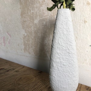 Könnte beinhalten: Eine weiße Keramikvase mit strukturierter Oberfläche. Die Vase ist hoch und schlank mit einem abgerundeten Boden. Sie ist leer und hat einen getrockneten Blumenstiel im Inneren.
