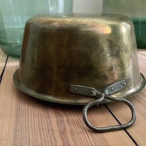 Op de afbeelding: Een vintage messing kom met een handvat. De kom is rond en heeft een glad, glanzend oppervlak. Het handvat is van metaal en is aan de zijkant van de kom bevestigd.