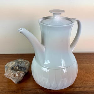 Peut inclure: Une cafetière en céramique blanche avec un couvercle et une poignée. La cafetière a un motif texturé sur le fond.