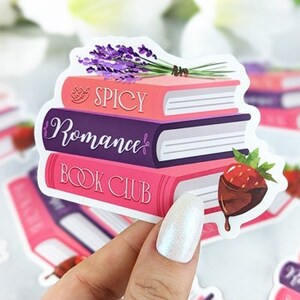 May include: A sticker featuring a stack of three books with the words "Spicy Romance Book Club" on the covers. The books are pink, purple, and pink, and there is a small strawberry with chocolate on top of the stack. The sticker is on a white background.