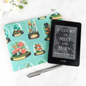 Puede incluir: Una funda de libro de tela azul turquesa con un dise&ntilde;o de setas y naturaleza. La funda est&aacute; decorada con ilustraciones de setas en c&uacute;pulas de cristal. El texto "Observe", "Torture" y "A Court of Mist and Fury" es visible en la funda. Un lector electr&oacute;nico Kindle est&aacute; sentado encima de la funda.