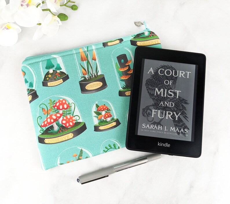 Puede incluir: Una funda de libro de tela azul turquesa con un dise&ntilde;o de setas y naturaleza. La funda est&aacute; decorada con ilustraciones de setas en c&uacute;pulas de cristal. El texto "Observe", "Torture" y "A Court of Mist and Fury" es visible en la funda. Un lector electr&oacute;nico Kindle est&aacute; sentado encima de la funda.