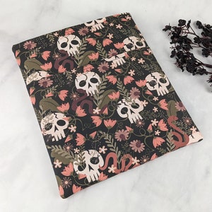 K&ouml;nnte beinhalten: Ein dunkler Stoff mit einem sich wiederholenden Muster aus wei&szlig;en Totenk&ouml;pfen, rosa Blumen und braunen Schlangen. Das Design umfasst auch gr&uuml;ne Bl&auml;tter und kleine wei&szlig;e Bl&uuml;ten. Der Stoff ist gefaltet, was auf eine Verwendung f&uuml;r einen Buchumschlag hindeutet.