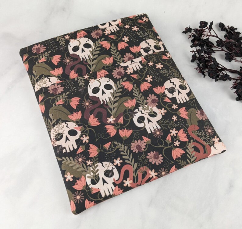 K&ouml;nnte beinhalten: Ein dunkler Stoff mit einem sich wiederholenden Muster aus wei&szlig;en Totenk&ouml;pfen, rosa Blumen und braunen Schlangen. Das Design umfasst auch gr&uuml;ne Bl&auml;tter und kleine wei&szlig;e Bl&uuml;ten. Der Stoff ist gefaltet, was auf eine Verwendung f&uuml;r einen Buchumschlag hindeutet.