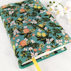 Könnte beinhalten: Ein türkisfarbener Buchumschlag mit einem Blumenmuster mit weißen Blüten, grünen Blättern und gelben und orangen Zitrusfrüchten.