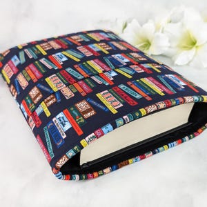 Könnte beinhalten: Eine marineblaue Buchhülle mit einem farbenfrohen Muster aus gestapelten Büchern. Die Buchhülle ist geöffnet und zeigt die Seiten eines Buches im Inneren. Die Buchhülle ist aus Stoff und hat ein schwarzes Futter.