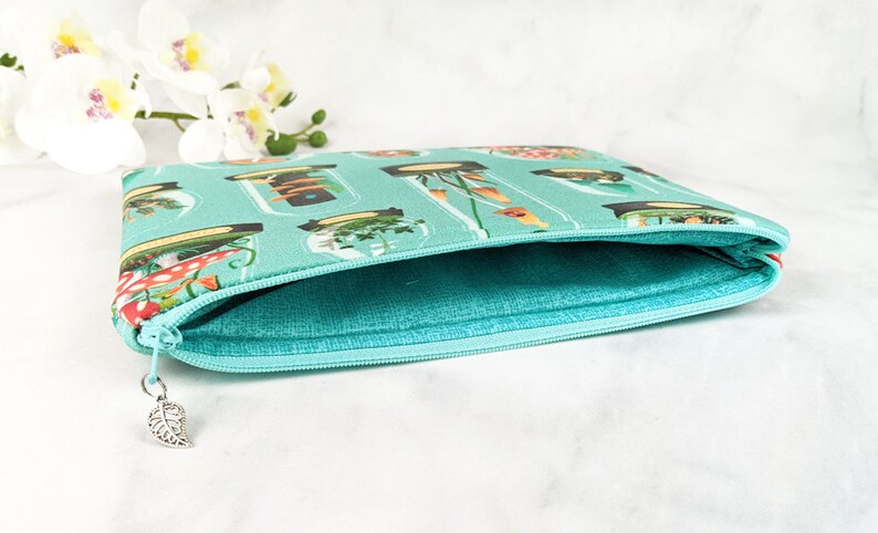 Puede incluir: Una bolsa de cremallera de color turquesa con un estampado floral que presenta frascos de plantas y hongos. La bolsa est&aacute; abierta y el forro es de color turquesa claro.