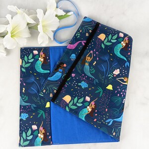May include: A blue fabric book cover with a mermaid and sea life pattern. The cover has a black elastic closure and a blue fabric lining.