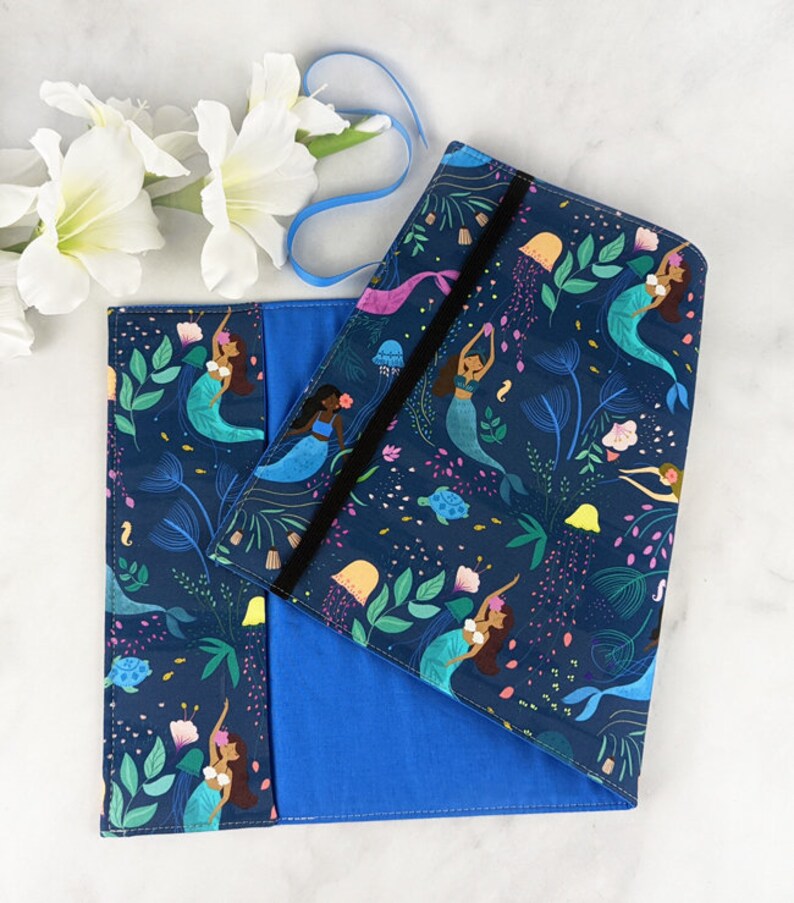 May include: A blue fabric book cover with a mermaid and sea life pattern. The cover has a black elastic closure and a blue fabric lining.