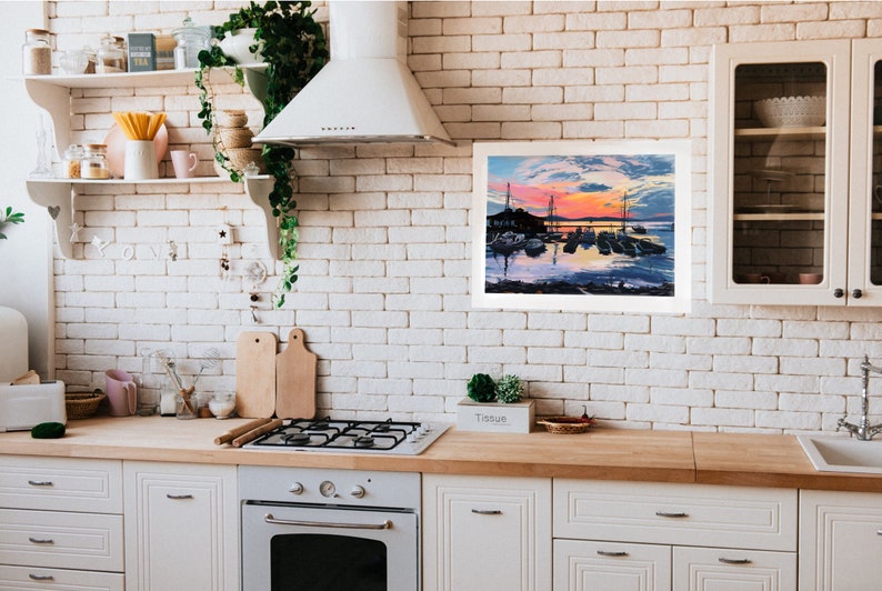 May include: A framed painting of a harbor scene with boats at sunset. The painting is hanging on a white brick wall in a kitchen.